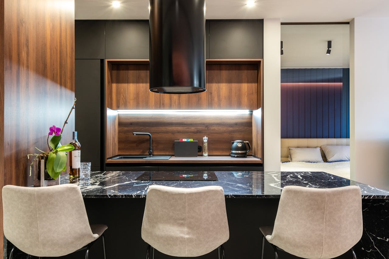 Contemporary kitchen with hob under hood against electric appliances and tap in light house with chairs